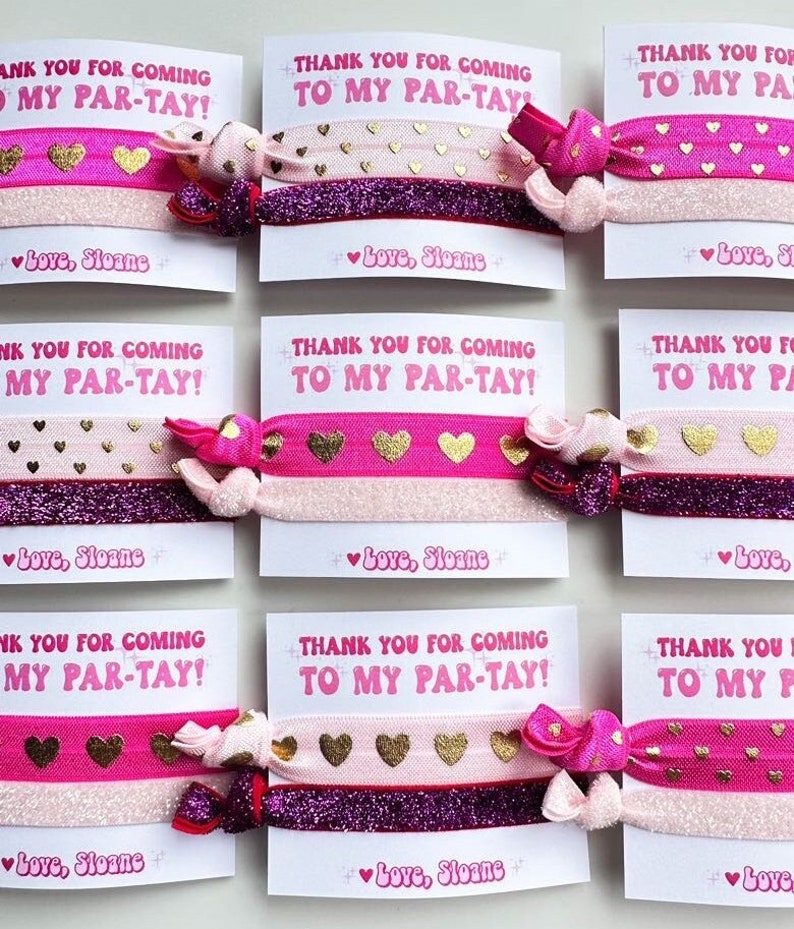 Pode incluir: El&aacute;sticos de cabelo rosa e dourados com motivos de cora&ccedil;&otilde;es, apresentados com cart&otilde;es brancos com o texto "THANK YOU FOR COMING TO MY PAR-TAY!". Cada el&aacute;stico tem um la&ccedil;o decorativo. O texto "Love, Sloane" tamb&eacute;m est&aacute; presente.