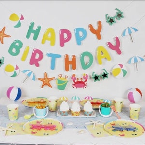 May include: A colorful "Happy Birthday" banner with beach-themed decorations, including starfish, beach balls, sunglasses, and a bucket and spade. The banner is hanging above a table set with plates, cups, and napkins featuring a similar beach theme.