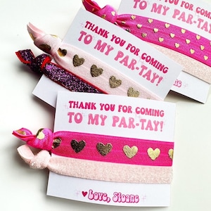 Pode incluir: Cart&otilde;es de lembrancinhas de festa com el&aacute;sticos de cabelo. Os cart&otilde;es dizem "THANK YOU FOR COMING TO MY PAR-TAY!" em rosa. Os el&aacute;sticos de cabelo s&atilde;o rosa, dourado e roxo brilhante, com detalhes de cora&ccedil;&atilde;o dourado. O cart&atilde;o inferior diz "Love, Sloane".