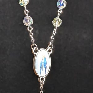 May include: A silver rosary necklace with iridescent crystal beads and a cross pendant. The oval centerpiece features an image of the Virgin Mary. The cross is detailed with blue, red, and green enamel.