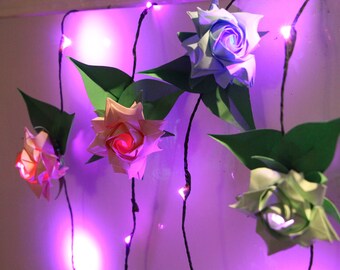 May include: A string of purple fairy lights with four origami paper roses hanging from them. The roses are in shades of pink, blue, and green.
