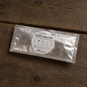 Op de afbeelding: Een zilveren zakje met ruwe lanoline, een pure wolvet, ligt op een houten oppervlak. Het zakje heeft een rond etiket met tekst en pictogrammen, waaronder "nz naturally" en "RAW LANOLIN". Het product is ongefilterd en weegt 100g.