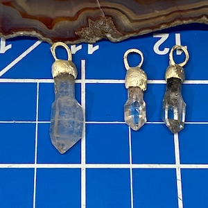 May include: Three clear quartz crystal pendants with gold-toned jump rings. The pendants are different sizes and are arranged in a row on a blue grid.