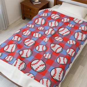 May include: A baseball-themed blanket with a repeating pattern of baseballs on a red and blue plaid background. The baseballs are white with red stitching and black outlines. The blanket is on a bed with a white pillow.