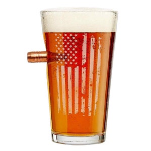 May include: A clear glass pint glass with a distressed American flag design etched into the side. The glass is filled with a light brown beer and has a copper bullet embedded in the side.