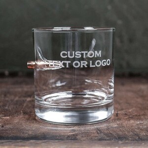 Puede incluir: Un vaso de vidrio transparente con una bala incrustada en el lateral. El vaso tiene las palabras "CUSTOM TEXT OR LOGO" grabadas en el lateral.