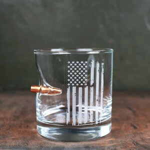 Puede incluir: Un vaso de vidrio transparente con un dise&ntilde;o de bandera estadounidense desgastado grabado en el lateral. Una bala de color cobre est&aacute; incrustada en el lateral del vaso.