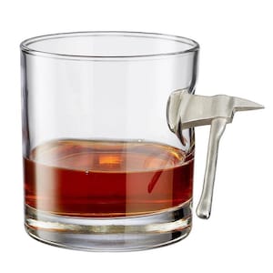 May include: A clear glass tumbler with a silver axe handle attached to the side. The glass is filled with a dark amber liquid.