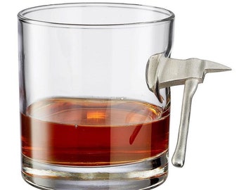 May include: A clear glass tumbler with a silver axe handle attached to the side. The glass is filled with a dark amber liquid.