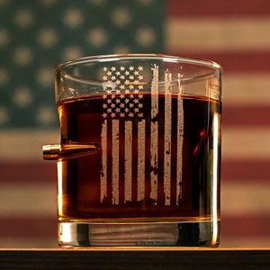 Puede incluir: Un vaso de whisky de vidrio transparente con una base con forma de bala. El vaso est&aacute; grabado con un dise&ntilde;o de bandera estadounidense desgastado. El vaso est&aacute; lleno de l&iacute;quido de color &aacute;mbar.