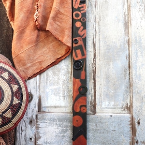 May include: A long, black wooden decorative piece with orange geometric shapes and circular accents. The piece is adorned with black metal rings and a natural fiber tie. A woven basket and orange fabric are in the background.