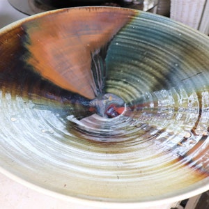 May include: A large, ceramic bowl with a swirling, abstract design in shades of brown, blue, green, and white. The bowl has a wide, shallow shape and a smooth, glazed finish.