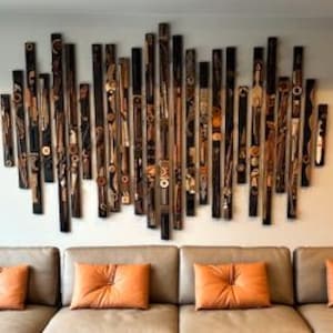 May include: Abstract wooden wall art composed of vertical, varying-length pieces in shades of brown and black. Some pieces feature circular and geometric designs. The artwork is mounted above a beige sofa with orange throw pillows.