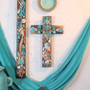 May include: Two wooden crosses decorated with turquoise and white seashells, coral, and driftwood. The crosses are hanging on a white wall with a turquoise fabric draped over the bottom.