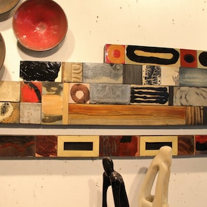May include: A wall art piece made of various ceramic tiles in different shapes and sizes. The tiles are arranged in a geometric pattern and feature abstract designs in shades of red, black, white, and brown.