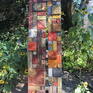 May include: Two vertical, rectangular mosaic art pieces. The pieces are composed of various textured tiles in shades of red, gold, brown, and black. The artwork is outdoors, surrounded by greenery and sunflowers.