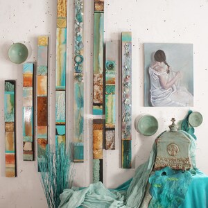 May include: A collection of vertical abstract art pieces in shades of blue, green, and brown, with a painting of a woman in a white dress on the wall behind them.
