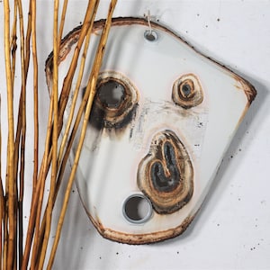 May include: A white ceramic wall hanging with a unique, abstract design featuring brown and black markings. The hanging has two holes for hanging and a rough, natural edge.