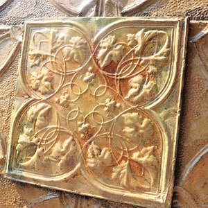 May include: A close-up of a gold-coloured metal tile with a decorative design of leaves and vines. The tile has a textured surface and is slightly worn.