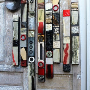 May include: A wall art piece made of wood and painted in black, white, red, and gold. The piece is made up of vertical strips of wood, each with a different pattern. The strips are arranged in a random order, creating a modern and abstract design.