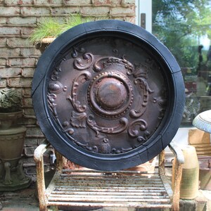 May include: A large, round, antique metal ceiling medallion with ornate embossed details. The medallion has a dark brown, aged appearance with a black outer rim. It rests on a vintage metal chair.