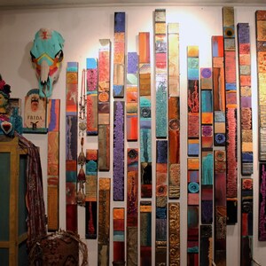 Peut inclure: Une installation murale d'art composée d'un ensemble de panneaux de bois texturés et colorés disposés verticalement. Les panneaux sont peints dans différentes nuances de bleu, vert, rouge, orange et marron, créant un design vibrant et abstrait.