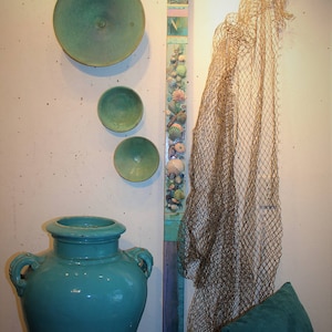 May include: A teal blue ceramic vase with two handles sits on a textured surface. A wooden panel with a collage of seashells and painted stripes is on the wall behind the vase. A brown fishing net hangs on the wall to the right of the vase.