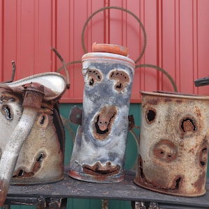 May include: Three rusty metal cans with holes cut out to create faces. The cans are arranged in a row on a wooden surface. The background is a red and green wall.