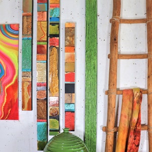 May include: A collection of colorful, abstract art pieces, including a painting with swirling lines of pink, yellow, and blue, and several vertical pieces made of wood and various materials. A wooden ladder with a fabric hanging on it and a small wooden sign with the word "Frida" on it are also visible.