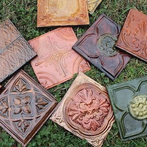 May include: Assortment of ten decorative ceramic tiles with various embossed patterns and colors, including green, brown, and gold. The tiles are arranged in a random pattern on a bed of green grass.