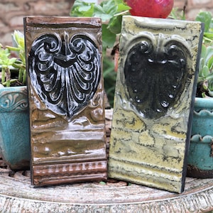 May include: Two decorative ceramic tiles with a heart-shaped design in black and a distressed finish. The tiles are brown and green with a rustic look.