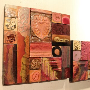 May include: A wall art piece made of small, colorful ceramic tiles. The tiles are arranged in a geometric pattern and feature various shapes, textures, and colors, including pink, red, brown, and gold.