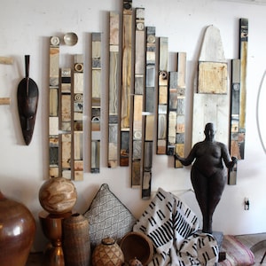 May include: A white wall with a collection of wooden and ceramic art pieces, including a sculpture of a nude figure. The art is arranged in a vertical pattern, with the pieces getting taller as they go up the wall. There are also a few decorative items on the floor, including a wooden bowl, a woven basket, and a patterned throw blanket.