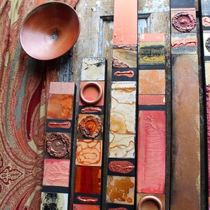Puede incluir: Una pieza de arte de pared hecha de madera con una variedad de texturas y colores. La pieza está compuesta por varios paneles verticales, cada uno con un diseño diferente. Los paneles están pintados en tonos de naranja, rojo y marrón, y algunos tienen un acabado metálico. La pieza es un diseño moderno y abstracto.