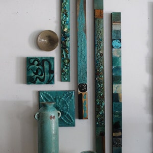 Puede incluir: Una exposición de pared de azulejos de cerámica de color verde azulado y turquesa y paneles de madera. Los azulejos están dispuestos en un patrón vertical y presentan varios diseños, que incluyen formas geométricas, patrones abstractos y un diseño en espiral. Dos jarrones de cerámica verde azulado están sobre un pedestal blanco.