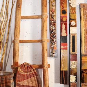 Könnte beinhalten: Eine Holzleiter lehnt an einer weißen Wand, an der ein geflochtener Korb am untersten Spross hängt. An der Wand sind vier vertikale Kunstwerke mit verschiedenen Materialien und Texturen ausgestellt.