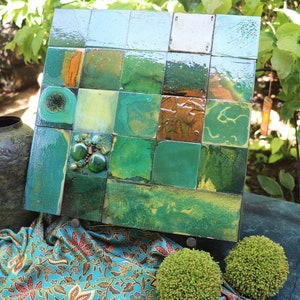 May include: A mosaic wall art piece featuring a grid of green and brown tiles with a glossy finish. The artwork is displayed outdoors, with a decorative vase and greenery in the background. The tiles are arranged in a square pattern.
