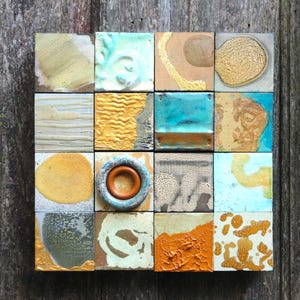 May include: A wall art piece made of 16 square ceramic tiles. The tiles are arranged in a grid pattern and feature various colors and textures, including blue, green, yellow, orange, and brown. Some tiles have a smooth surface, while others have a textured surface.