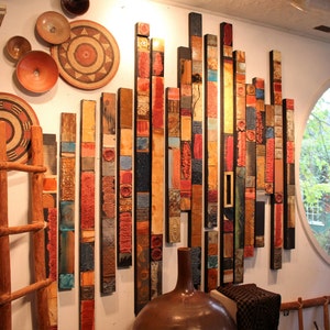 May include: A wall art piece made of colorful wooden slats, arranged in a vertical pattern. The slats are painted in various shades of red, orange, yellow, blue, and green. The piece is mounted on a white wall, and there are several decorative plates hanging on the wall to the left of the art piece.