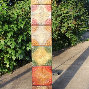 Puede incluir: Una pila de seis azulejos decorativos con un patrón repetido de dorado, verde, rojo y crema. Los azulejos están hechos de estaño y tienen un acabado desgastado.