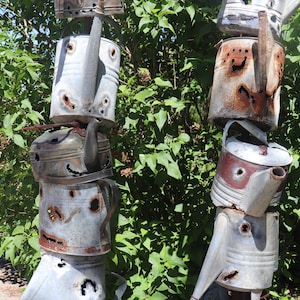 May include: Two tall sculptures made from stacked, rusty, metal watering cans. The cans have holes and are arranged in a totem pole style. The sculptures are in front of a green bush.