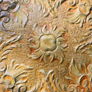 May include: A close-up of a decorative tile with a raised, ornate design. The tile is a warm, golden brown color with a distressed, aged look.