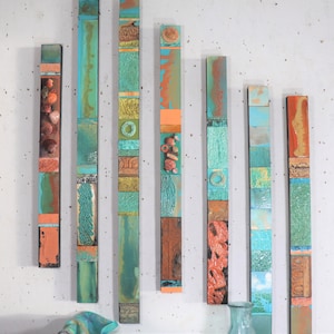 Può includere: Una collezione di sette opere d'arte murale verticali in legno. Ogni pezzo rettangolare presenta una varietà di colori, tra cui verde acqua, arancione e marrone. Alcuni pezzi presentano conchiglie e superfici strutturate, creando un'esposizione artistica unica.