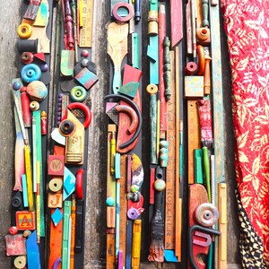 Può includere: Un'opera d'arte murale colorata realizzata con vari materiali riciclati, tra cui legno, metallo e plastica. L'opera presenta una varietà di colori, forme e texture, ed è disposta in uno schema verticale. L'opera è montata su uno sfondo in legno ed è circondata da un tessuto a motivi rossi e dorati.