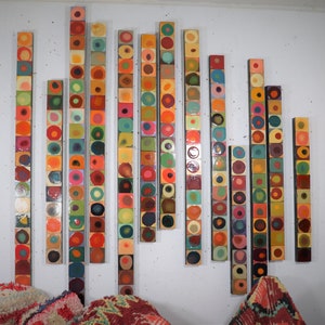 Op de afbeelding: Een wandinstallatie met kunst, bestaande uit een reeks verticale panelen, elk samengesteld uit kleine, kleurrijke, ronde tegels. De tegels zijn gerangschikt in een herhalend patroon, waardoor een visueel dynamisch en abstract ontwerp ontstaat.