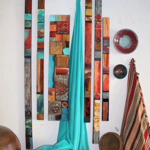Puede incluir: Una pieza de arte de pared hecha de azulejos coloridos dispuestos en tiras verticales. Los azulejos son en tonos de rojo, naranja, marrón, azul y verde. Una tela turquesa cuelga frente a la pieza de arte.