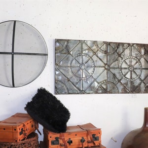 Op de afbeelding: Een witte muur met een rond metalen scherm, een rechthoekige metalen wanddecoratie met een geometrisch patroon, een zwart pluche kussen, twee bruine koffers, een rieten mand en een bruine keramische vaas.