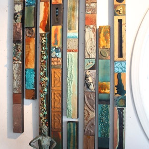 May include: A wall art installation made of multiple vertical strips of ceramic tiles. The tiles are arranged in a random pattern and feature various colours, textures, and designs. The tiles are primarily shades of blue, green, brown, and orange.