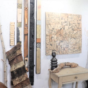 May include: A white wall with a variety of wall art, including a black and white abstract painting, a woven rug, and several wooden sculptures. There is also a small wooden table with a vase and a piece of driftwood on top.