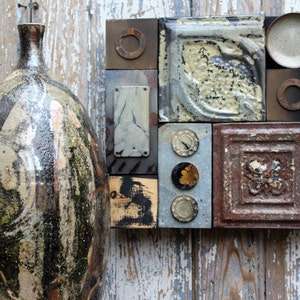 Może przedstawiać: Ceramiczny wazon z abstrakcyjnym wzorem w kolorze czarnym, białym i brązowym stoi obok ściennej dekoracji wykonanej z różnych metalowych płytek. Płytki są ułożone w siatkę i mają różne faktury i kolory, w tym rdzawo brązowy, srebrny i złoty.
